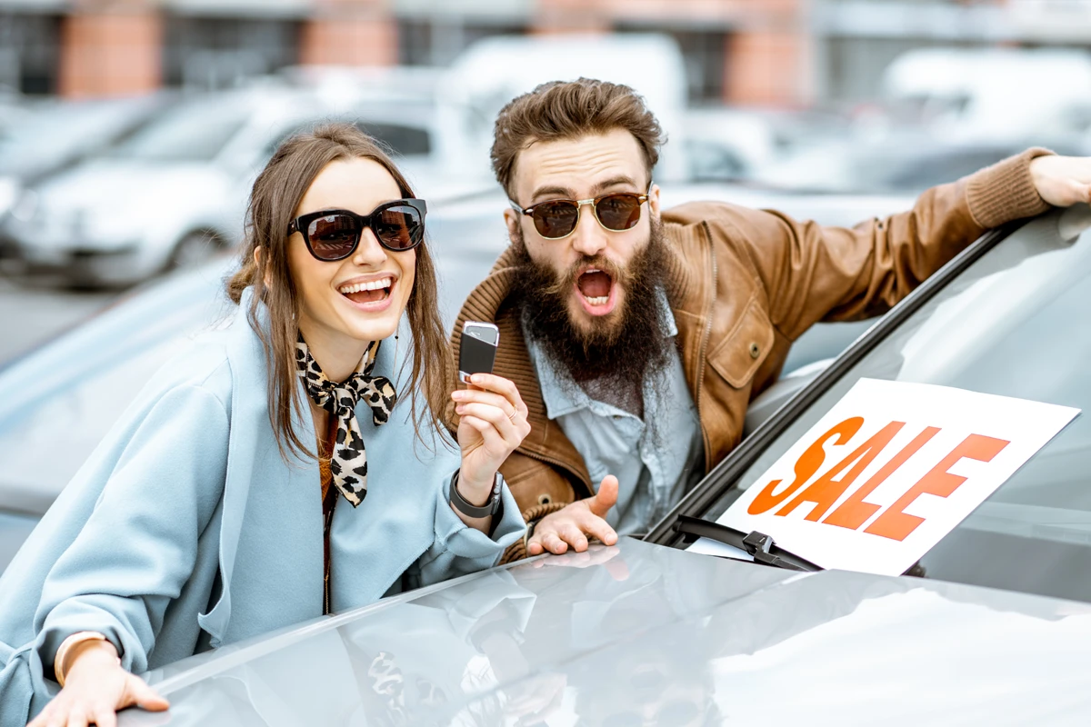 Excited couple with sale sign at used car dealership.