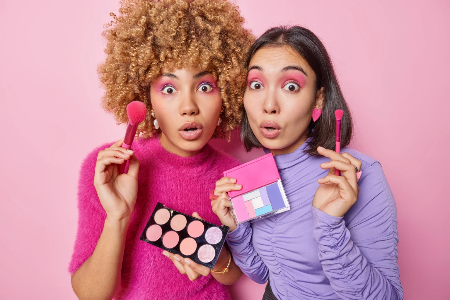 Two women applying makeup, surprised expressions, pink background