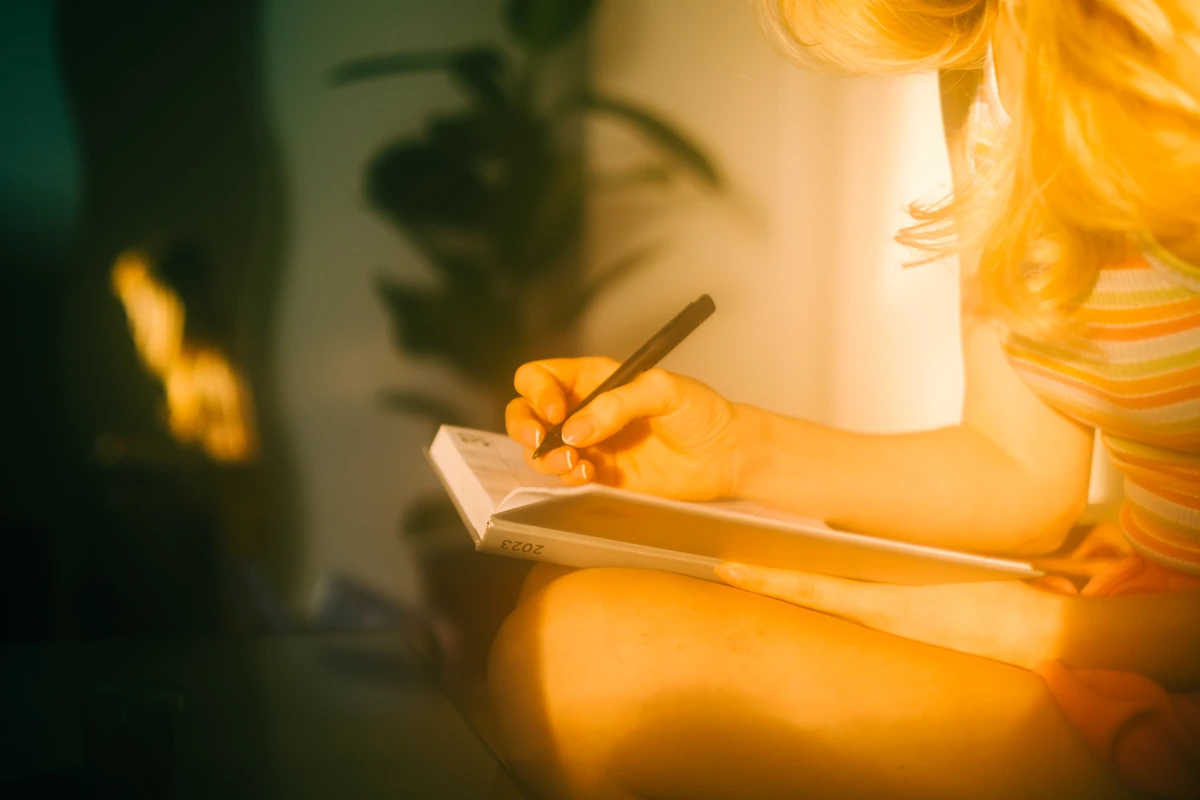 Woman writing in notebook beside window with plants.