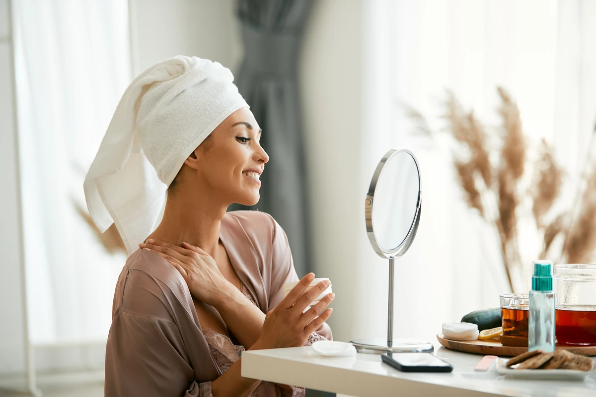 Woman in towel turban admiring reflection, skincare products nearby