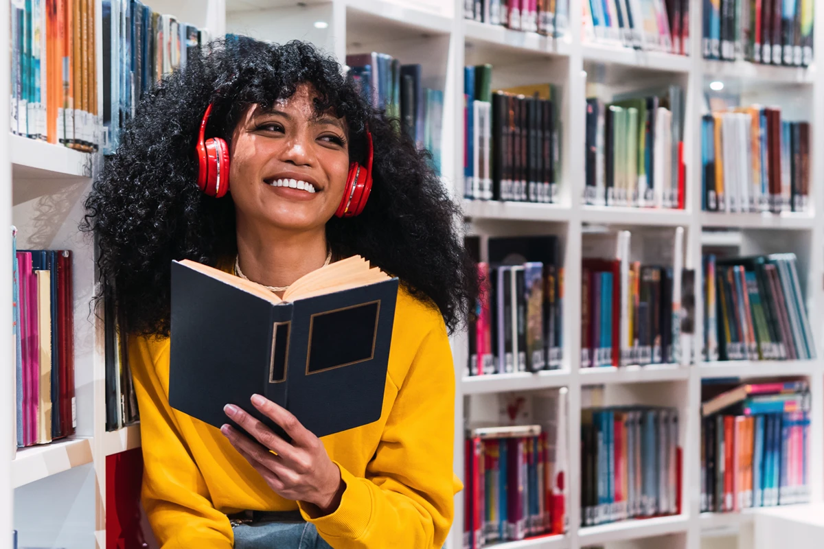 Joyful woman reading book with headphones in library