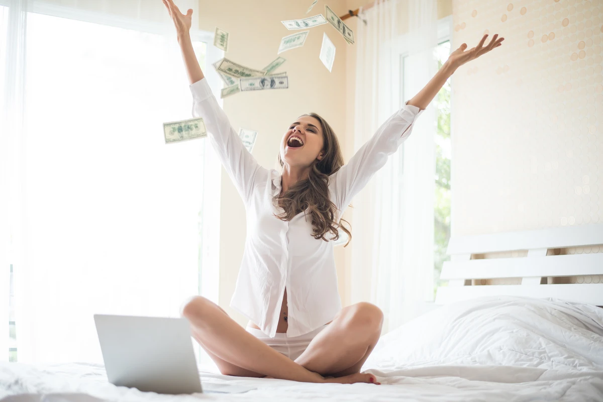 Woman celebrating with flying money sitting near laptop