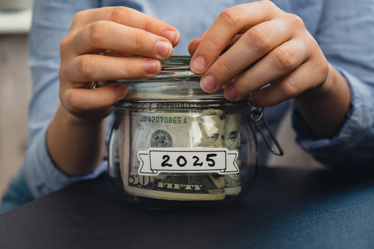 Hands closing a glass jar filled with cash labeled "2025" on a dark surface.