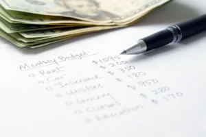 Monthly budget list with expenses and a stack of cash and a pen on a white background.