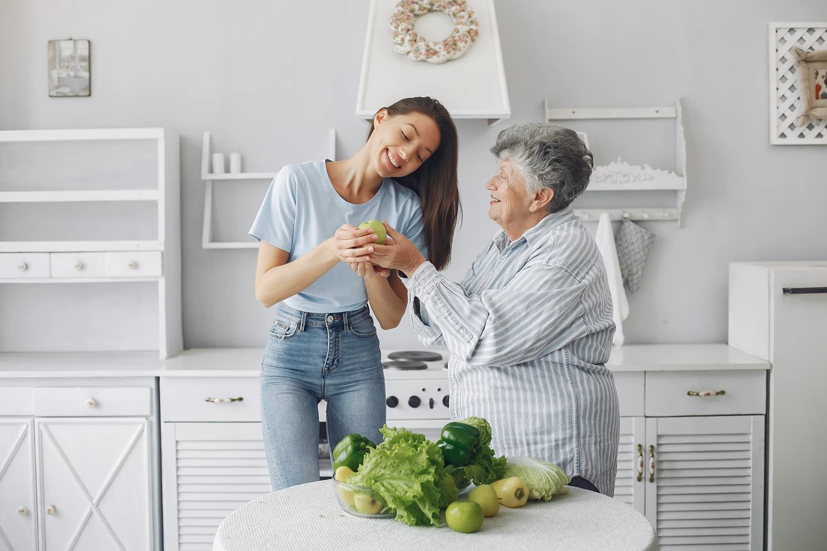 A young woman and an elderly woman share a joyful moment while handling fresh vegetables in a bright kitchen.