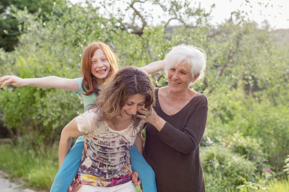 A grandmother, mother, and daughter share laughter outdoors, enjoying a playful moment in a lush garden.