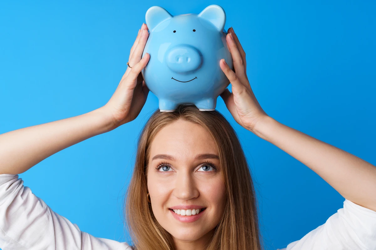 Woman smiling while balancing a blue piggy bank on her head against a bright blue background.