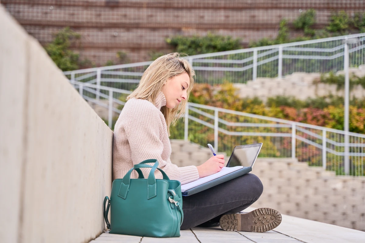 Woman sitting on a bench outdoors, writing in a notebook with a laptop beside her and a green handbag nearby.