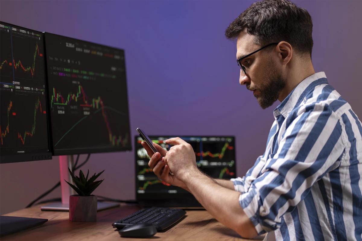 Man in a striped shirt analyzes stock market data on multiple screens while using a smartphone.