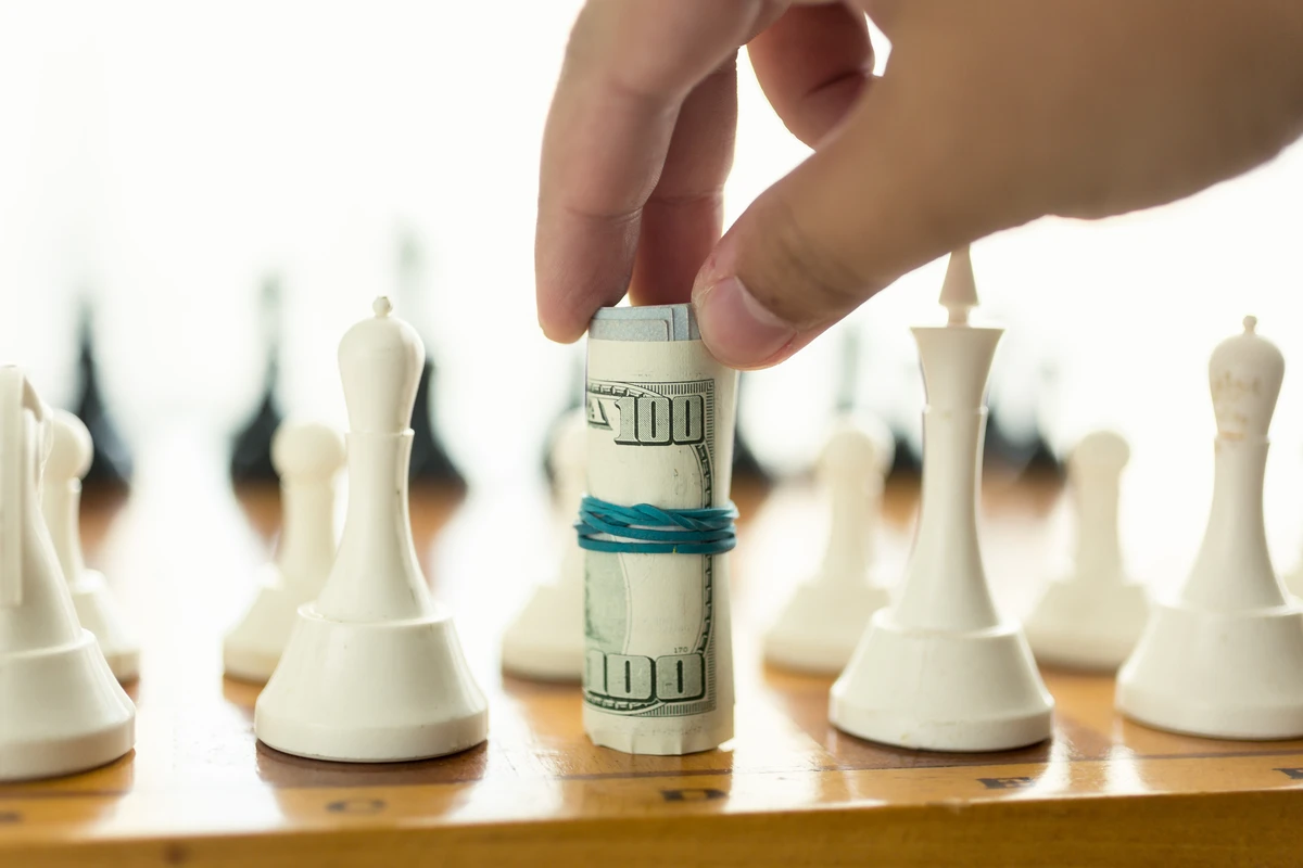 A hand places a rolled hundred-dollar bill among white chess pieces on a wooden board.
