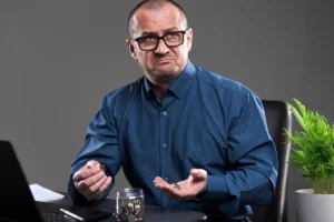 Man in a blue shirt gestures expressively while sitting at a desk with a laptop and a jar of coins.