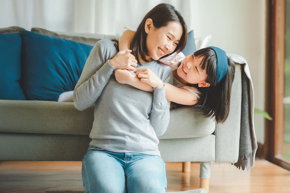 A mother and daughter share a joyful moment while sitting on a couch in a bright, cozy living room.