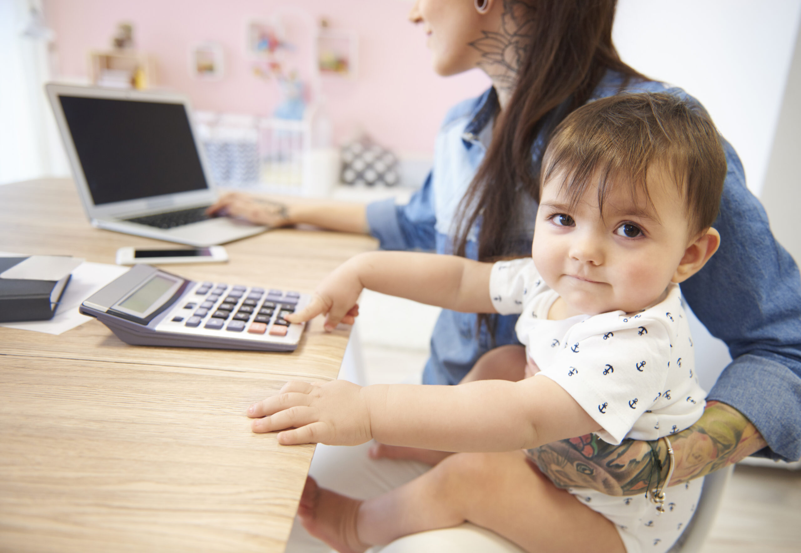 A toddler sits on a woman's lap, reaching for a calculator on a desk with a laptop and notebook nearby.