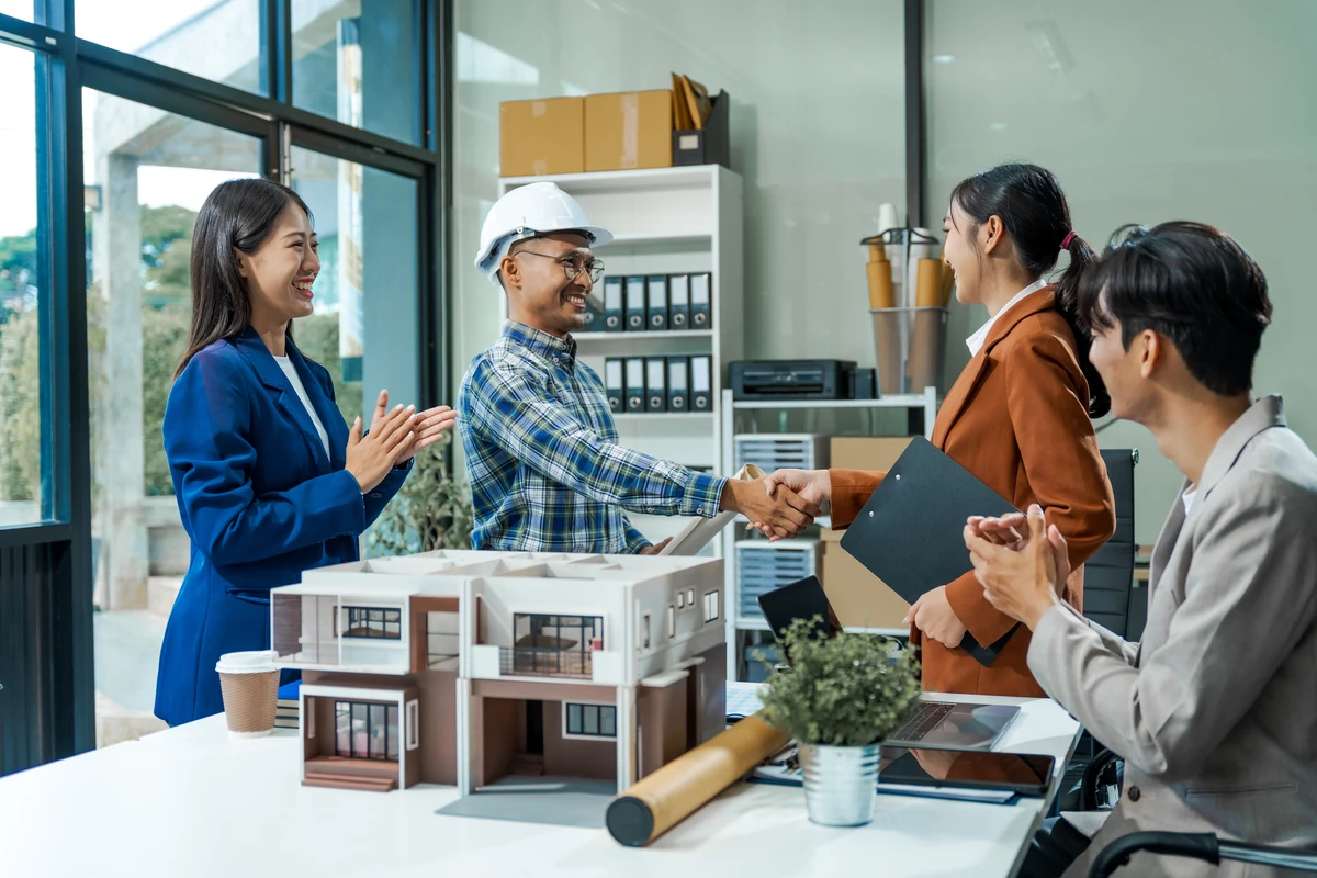 Team members celebrate a successful handshake in a modern office with architectural models on the table.