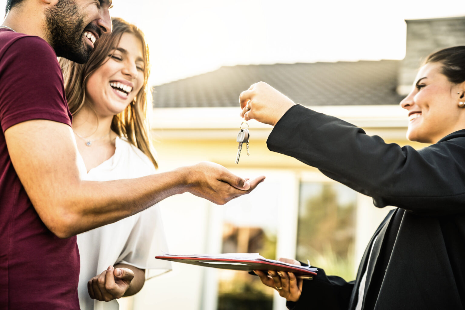 A woman hands house keys to a smiling couple outside a home, celebrating their new property.