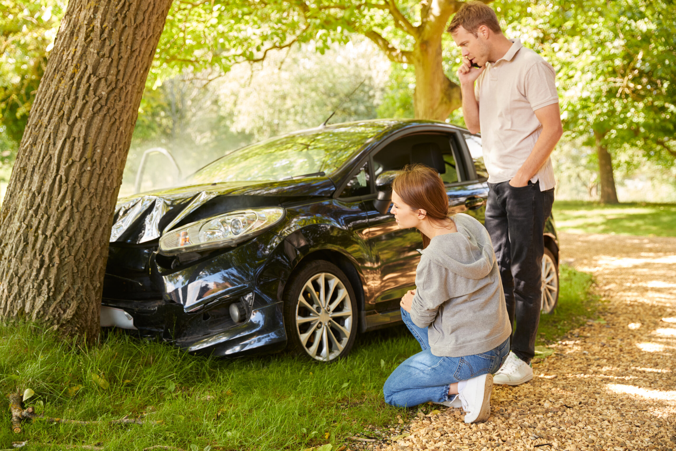 A man stands by a damaged car while a woman inspects the front near a tree in a sunny outdoor setting.