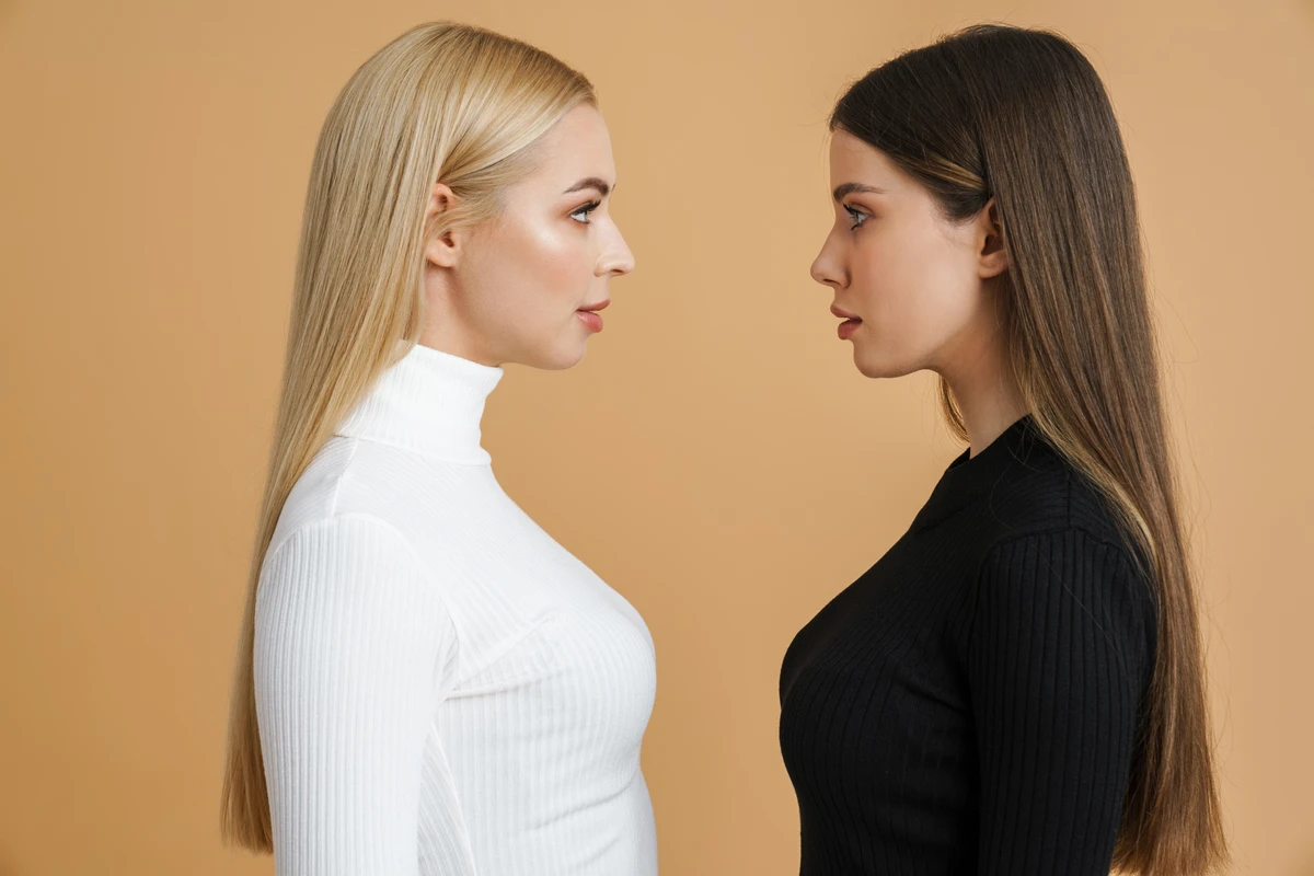 Two women with long hair face each other, one in a white turtleneck and the other in a black top, against a beige background.
