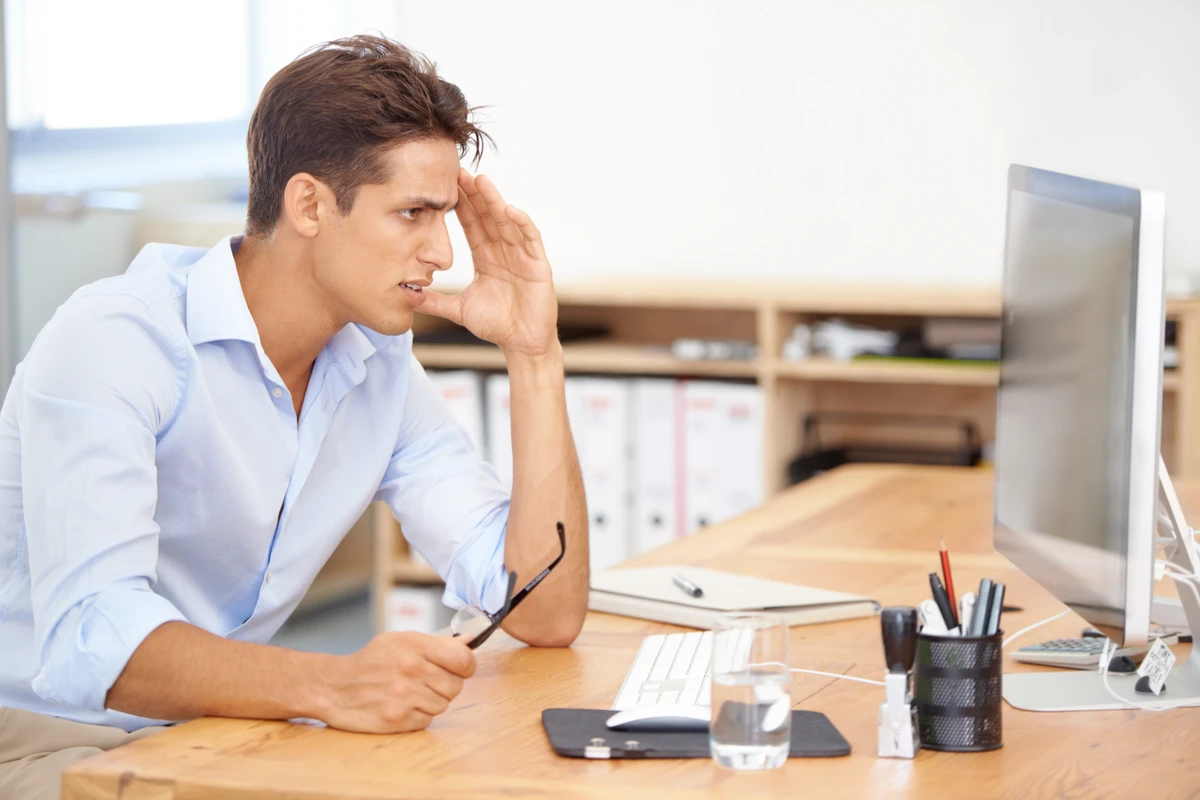 Frustrated man in a blue shirt sits at a desk, holding glasses while looking at a computer screen.