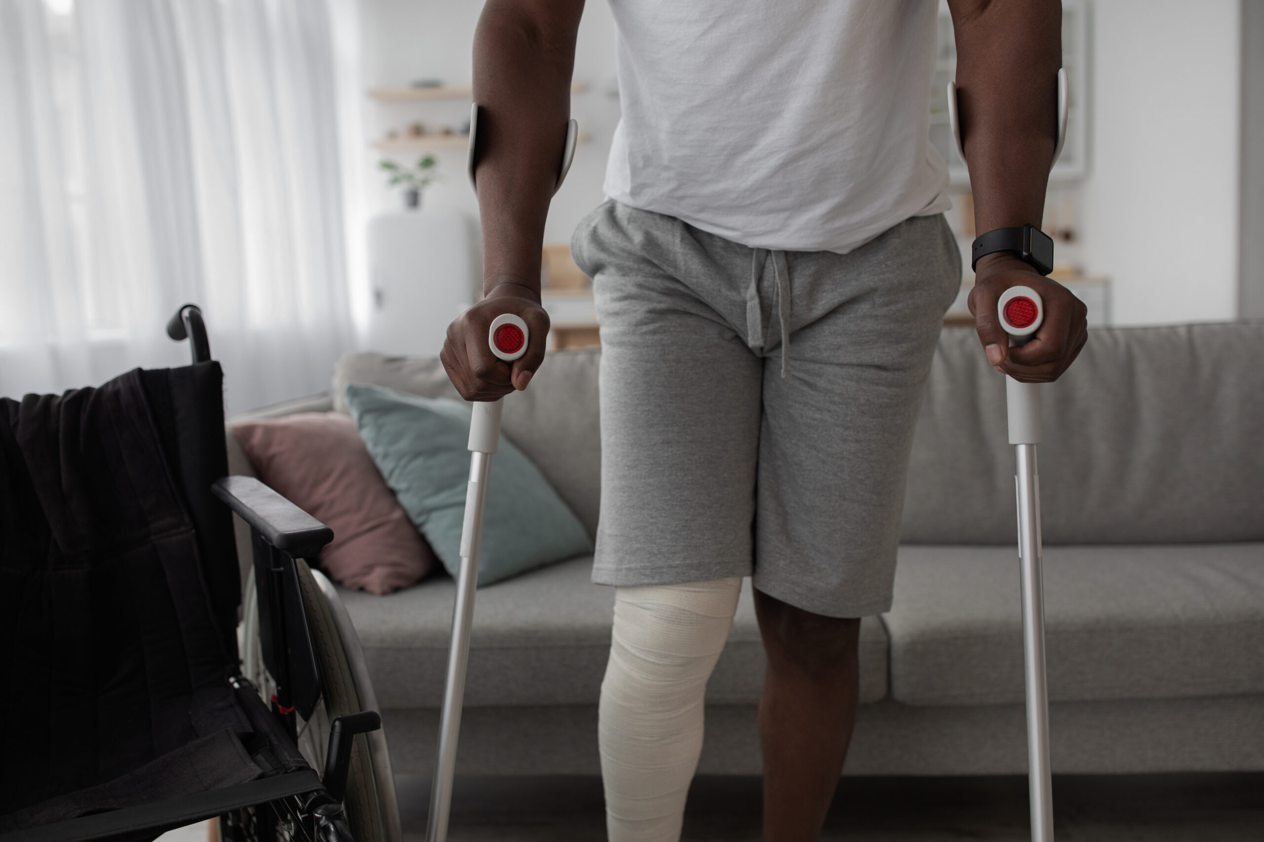Person using crutches to walk in a living room with a wheelchair and couch in the background.