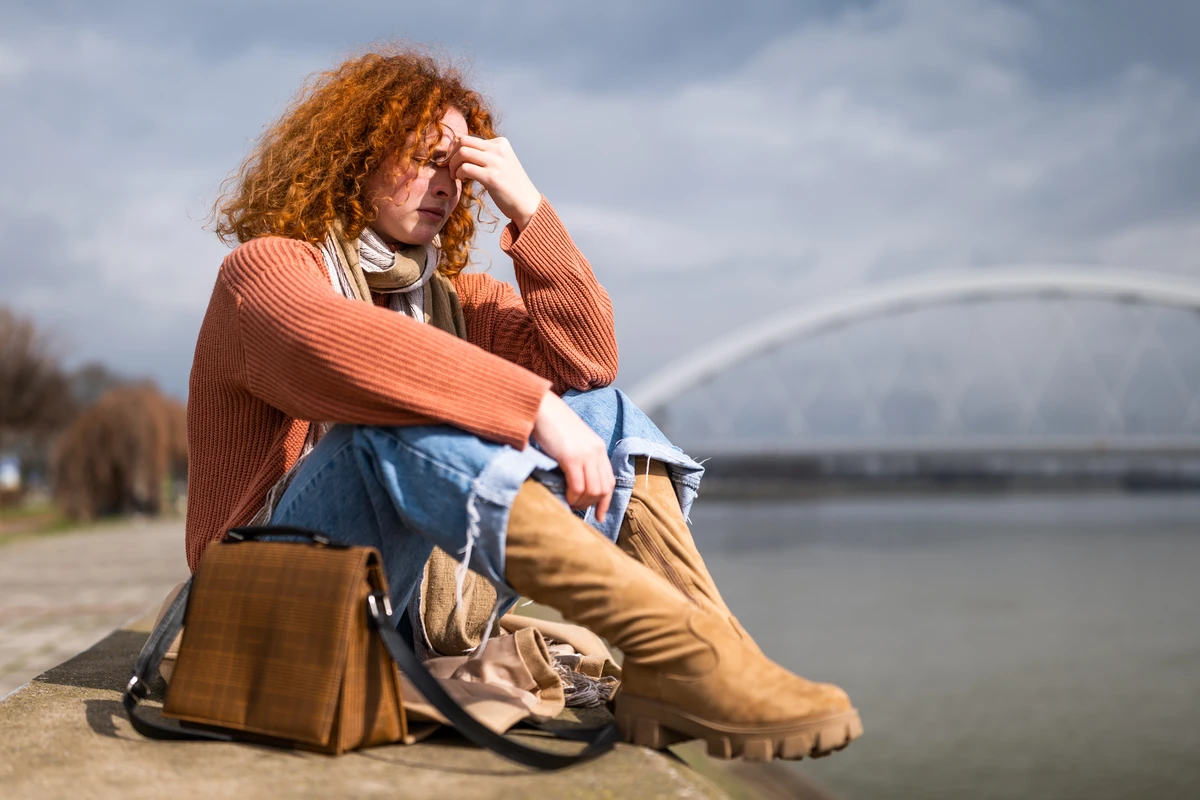 A woman with curly red hair sits by a river, looking pensive while wearing a cozy sweater and boots.