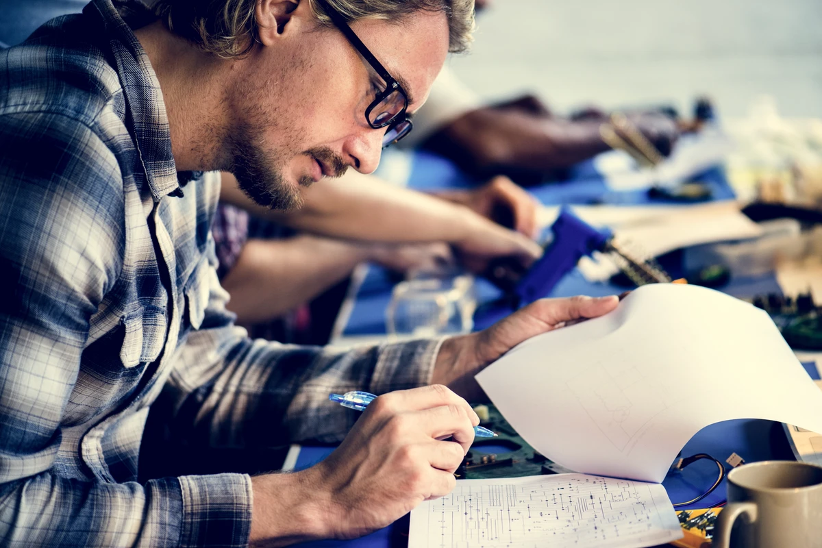 A man in glasses focuses on a blueprint while working on a project at a table with others.