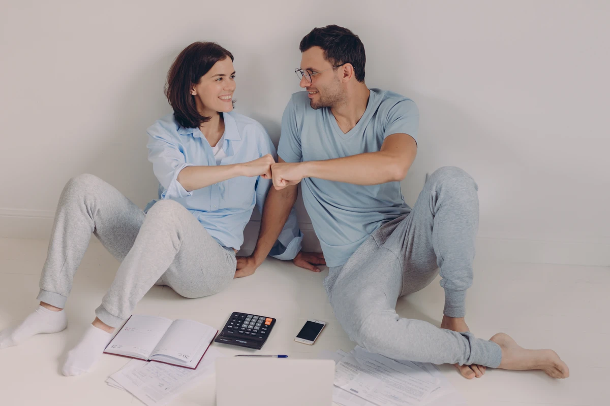 A couple sitting on the floor, smiling and fist-bumping, surrounded by paperwork and a calculator. financial boundaries