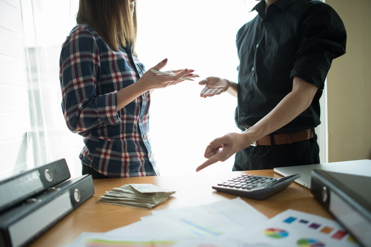 Two people discussing finances at a desk with cash and documents, while a calculator is visible.
