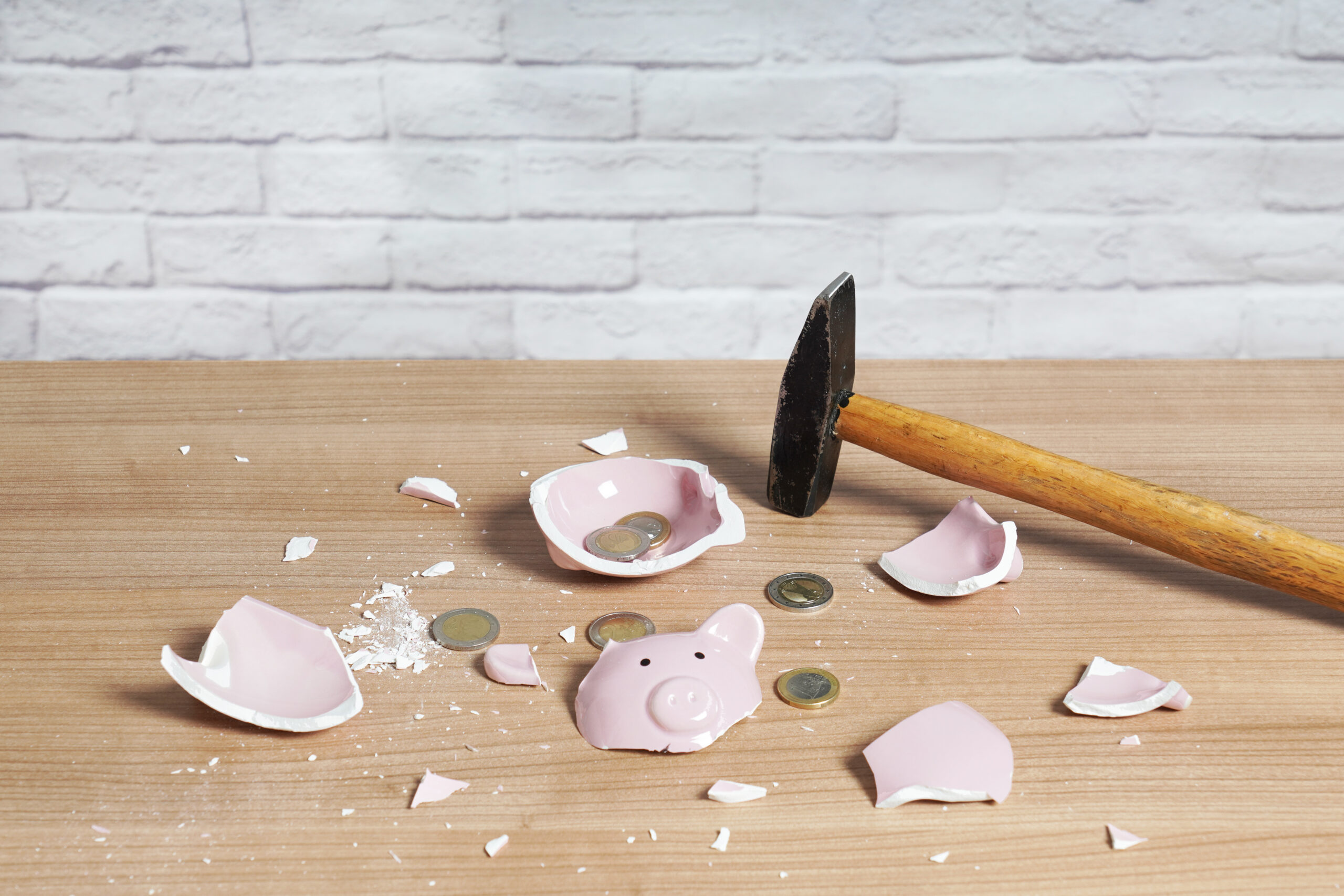 Broken pink piggy bank pieces scattered on a wooden table with coins and a hammer nearby.