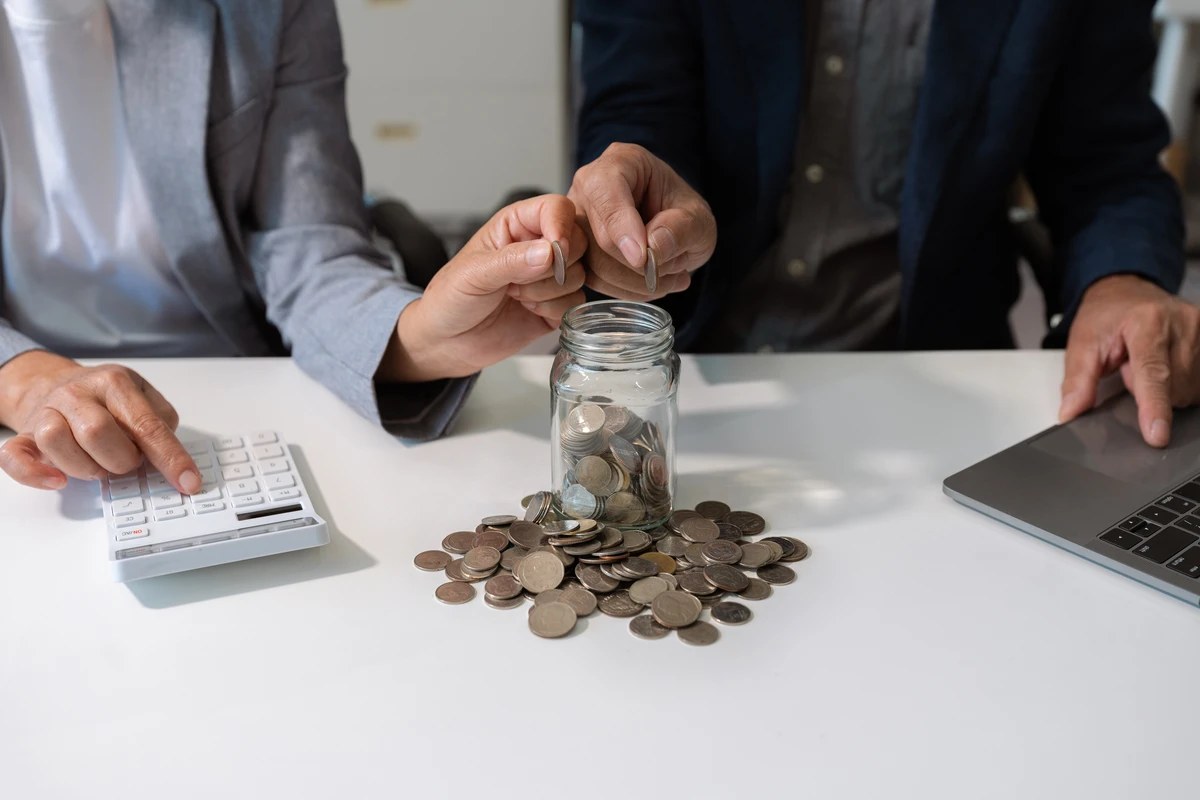 Two people adding coins to a jar while using a calculator and laptop on a desk.