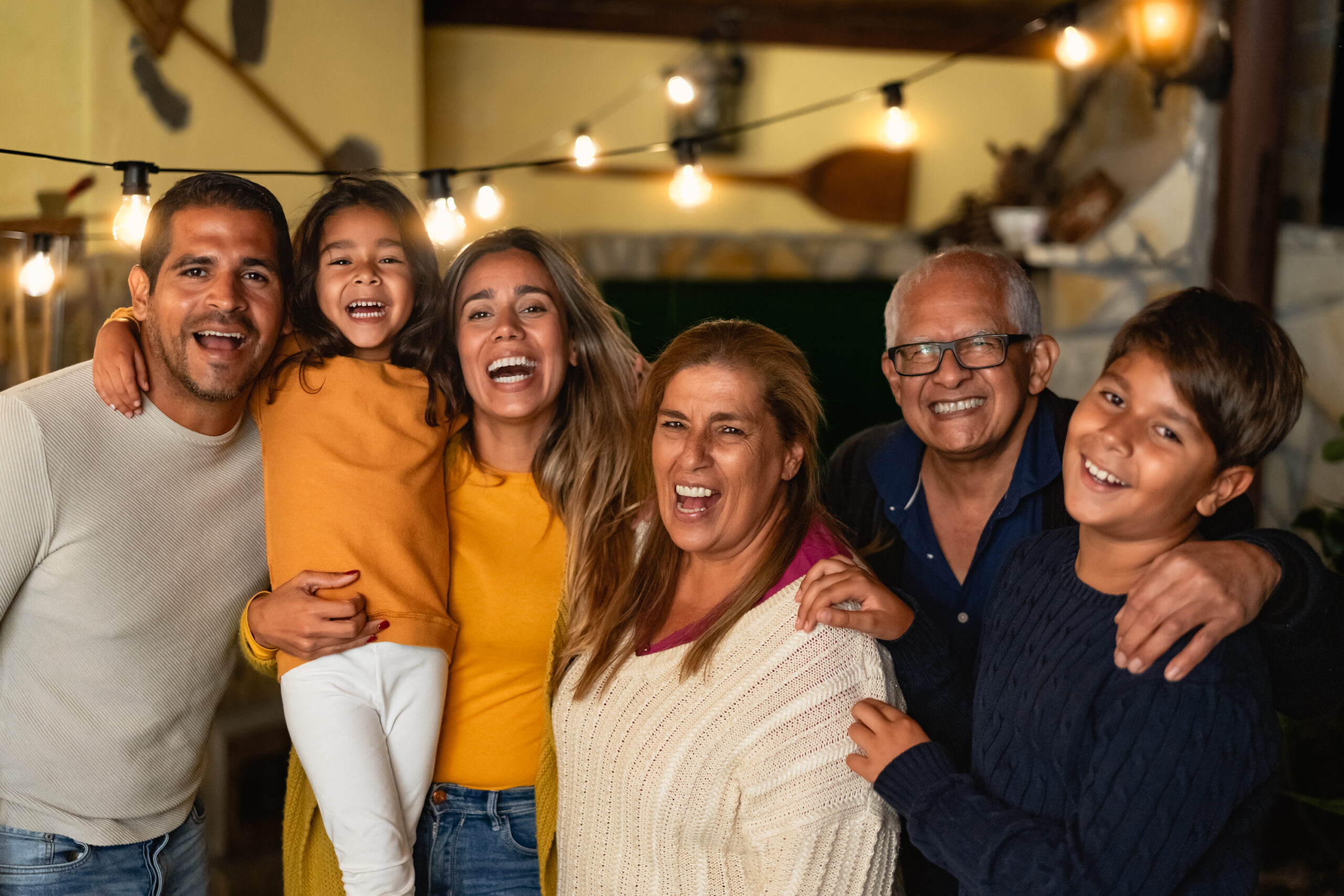 A joyful family of six smiles together under warm string lights in a cozy indoor setting.