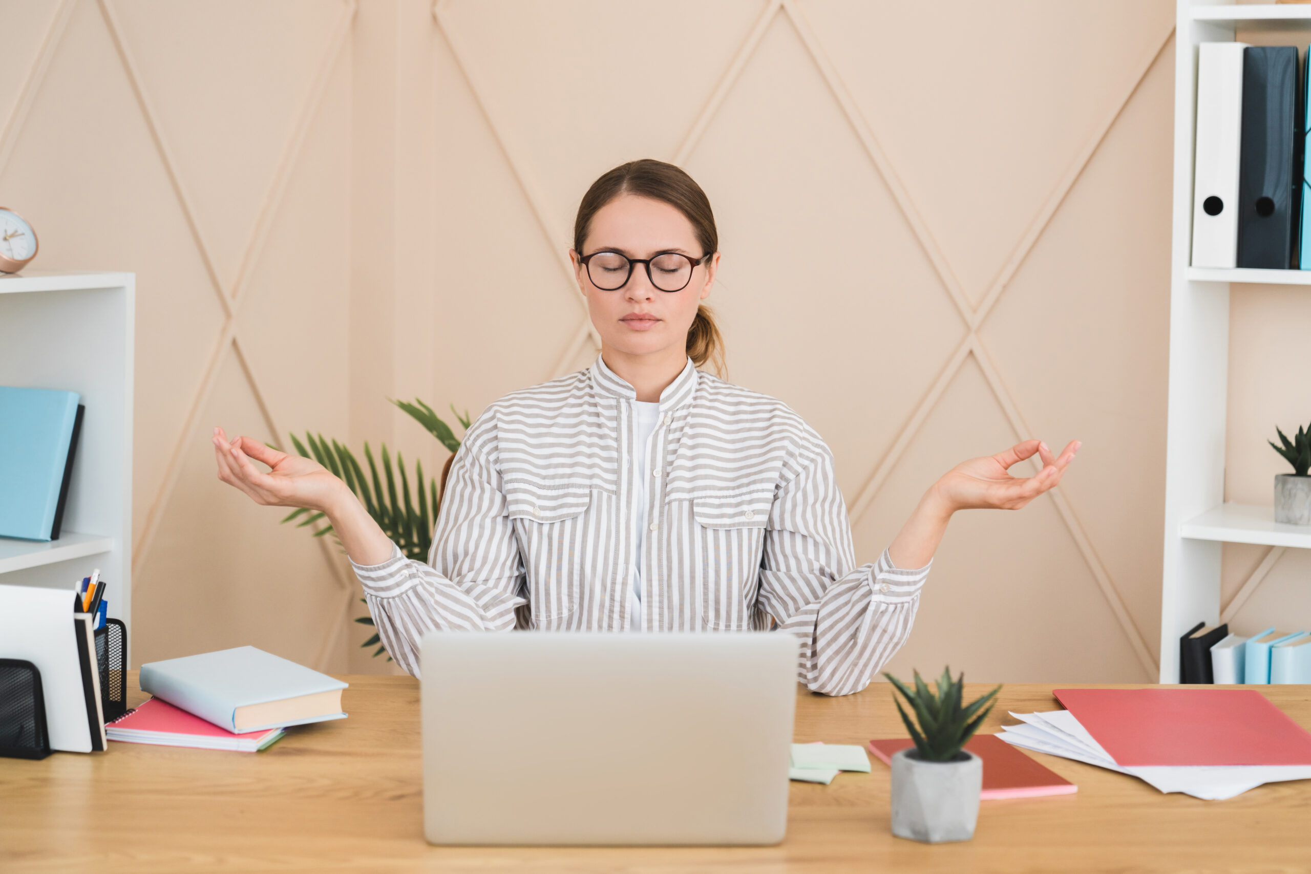 Woman meditating at a desk with a laptop, surrounded by books and office supplies in a calm workspace.