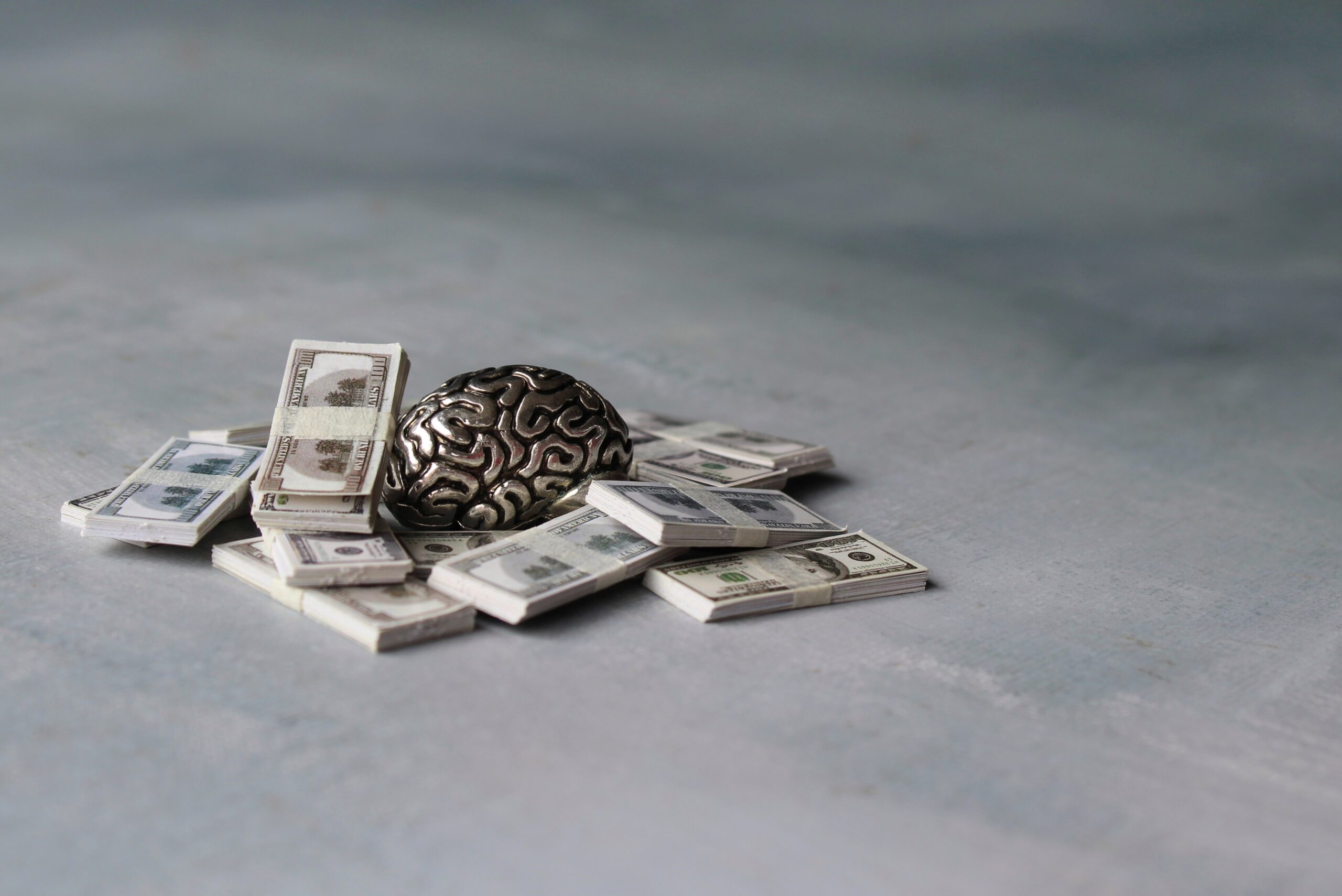 A silver brain sculpture rests on a pile of stacked hundred-dollar bills against a muted background. Changing your money mindset