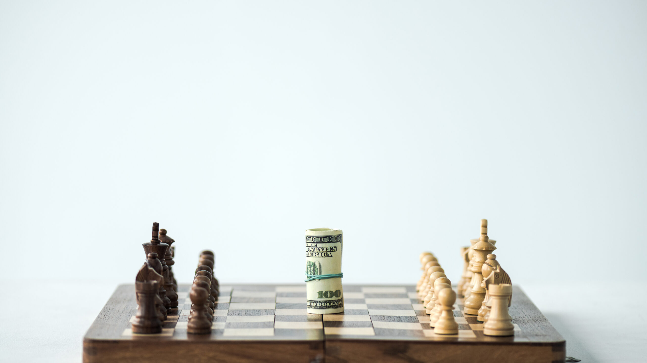 Chess pieces flank a rolled $100 bill positioned at the center of a chessboard.