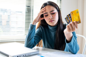 A young woman looks stressed while holding multiple credit cards at a desk with a laptop and paperwork. emergency fund