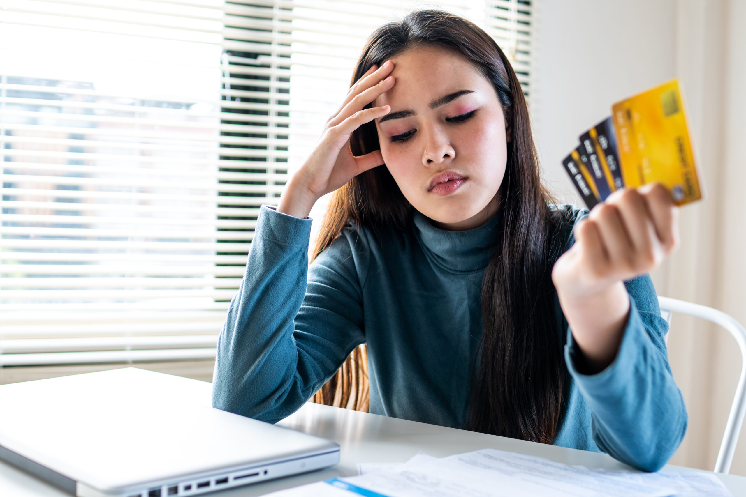 A young woman looks stressed while holding multiple credit cards at a desk with a laptop and paperwork. emergency fund