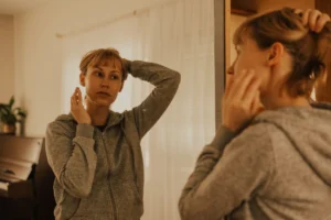 A woman in a gray hoodie examines her reflection while styling her hair in a softly lit room. Pretty privilege financial implications