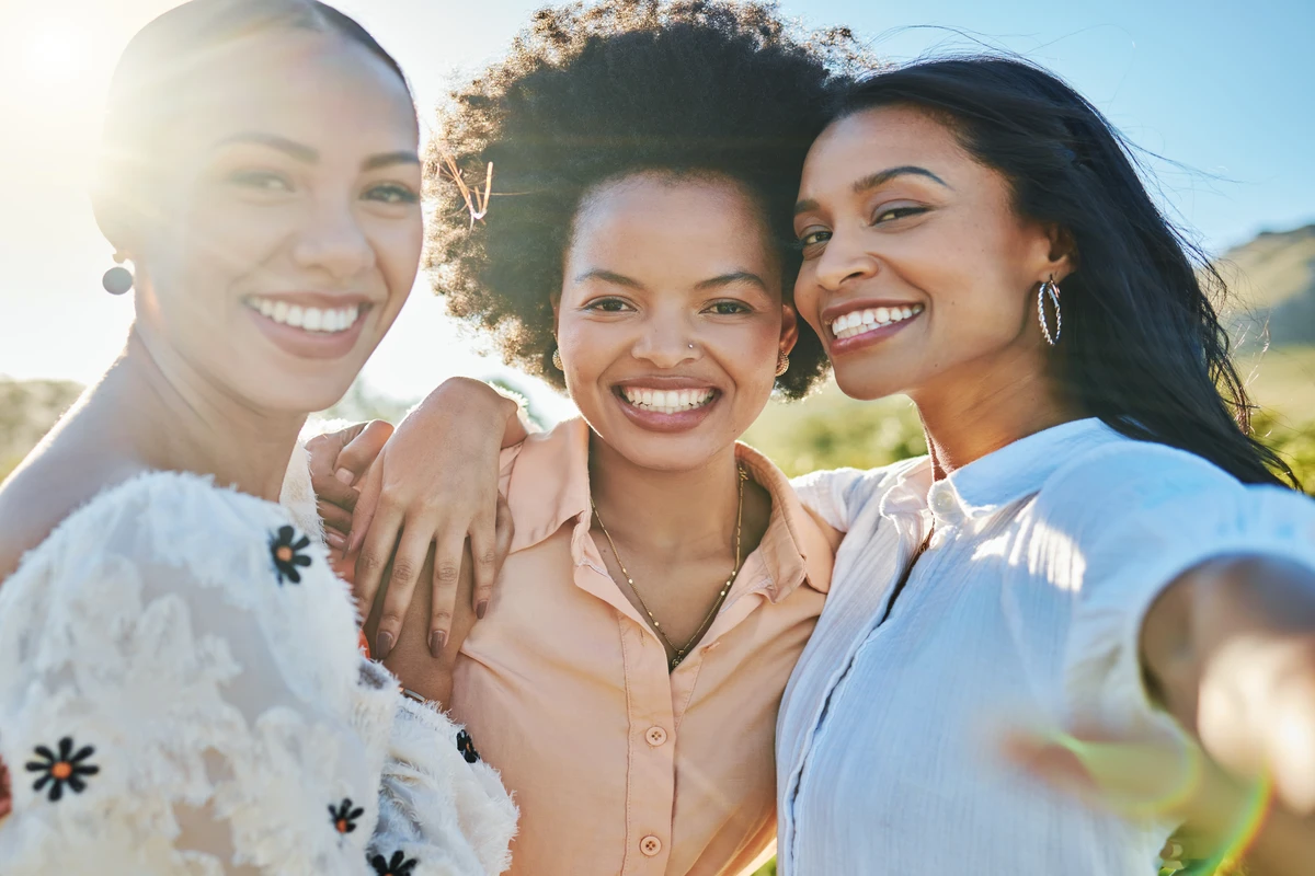Three smiling women pose together outdoors, enjoying a sunny day in a natural setting. pretty privilege financial implications
