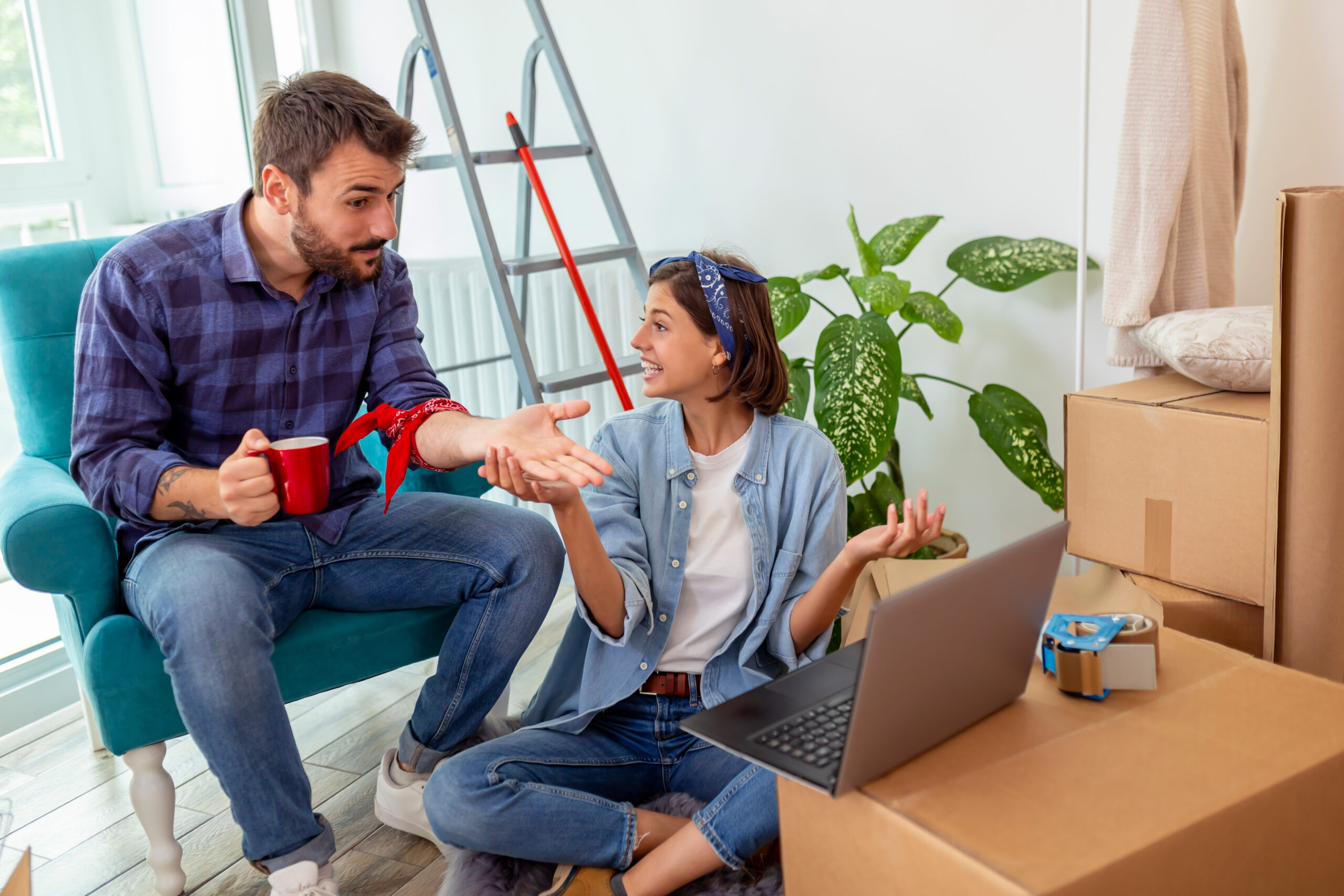 A man and woman discuss moving plans amidst unpacked boxes and a laptop in a bright, cozy room.