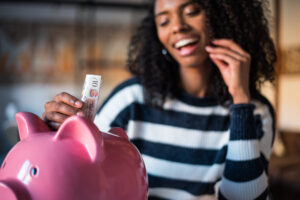 A woman joyfully deposits a £10 note into a pink piggy bank at home. saving strategies for important life events
