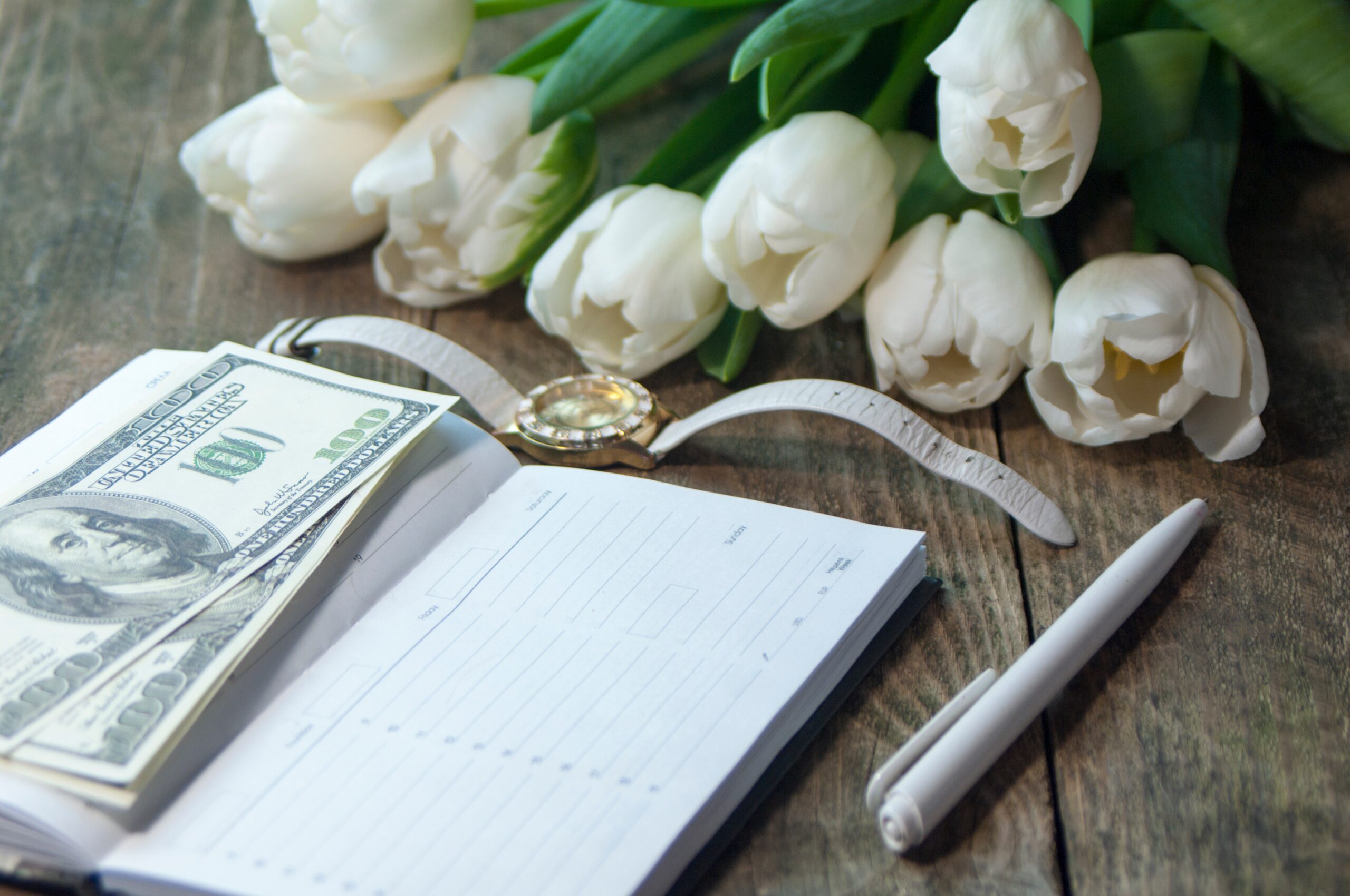 A hundred-dollar bill rests on an open planner beside a watch, pen, and white tulips on a wooden table.