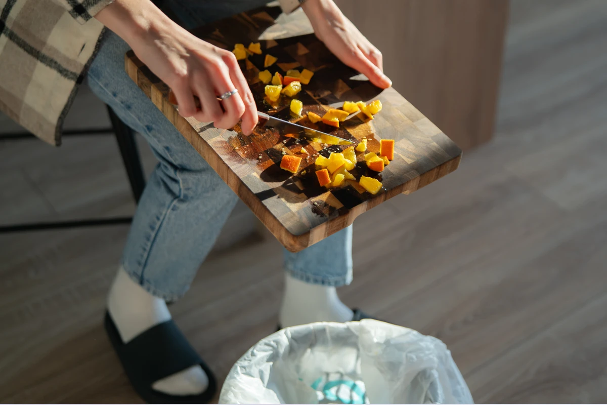 Person chopping colorful vegetables on a wooden cutting board, with a trash bin nearby.