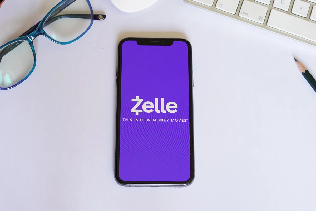 Zelle app displayed on a smartphone with glasses and a pencil on a desk.