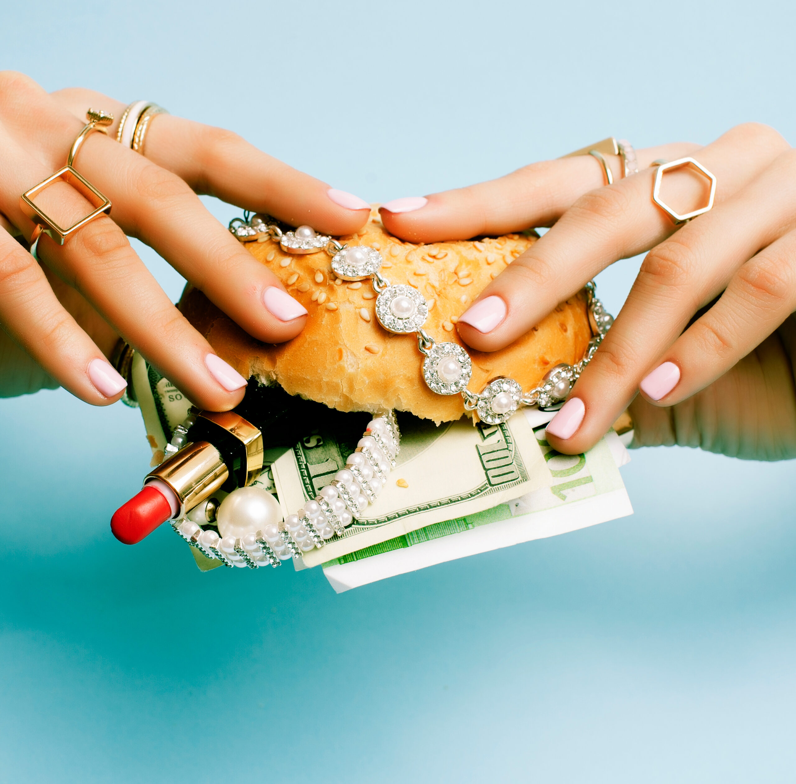 Hands adorned with rings hold a hamburger layered with cash, jewelry, and lipstick against a blue background.