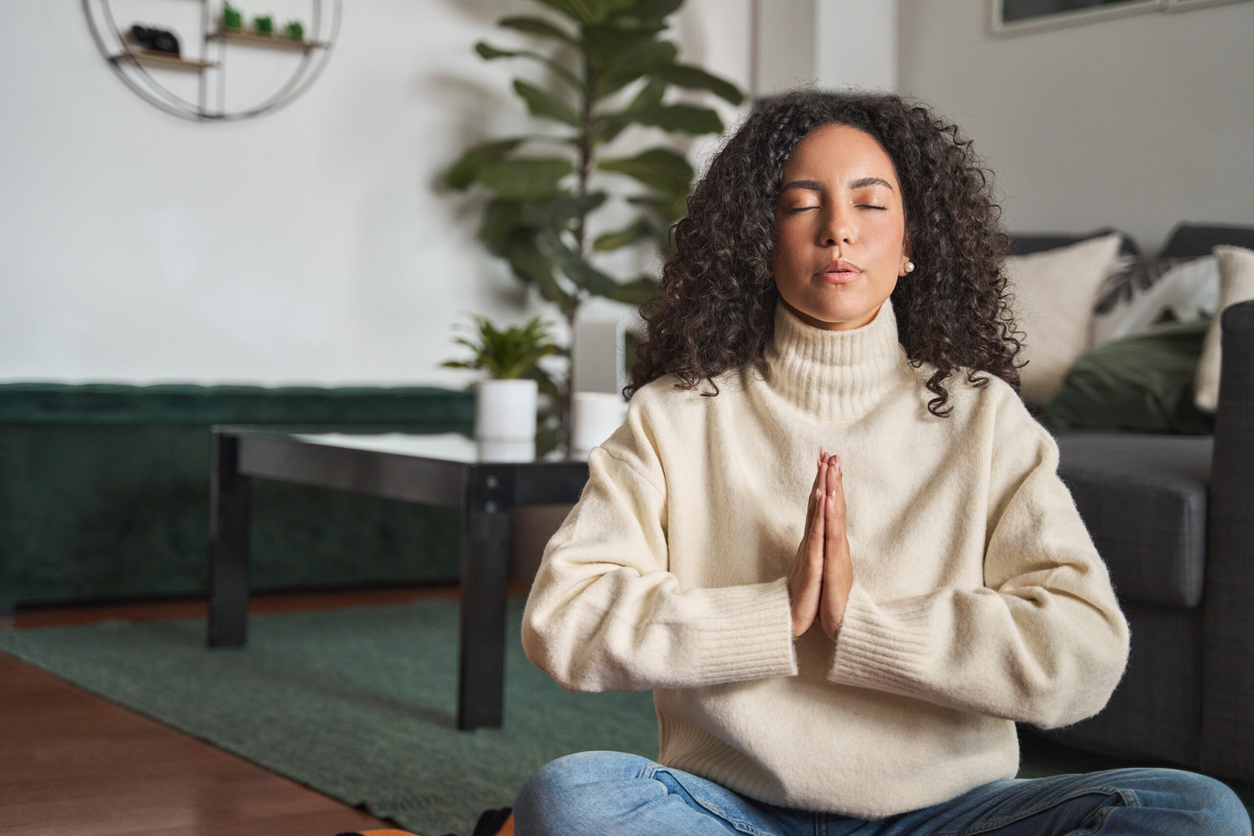 Woman meditating with hands in prayer position, surrounded by a cozy living room setting.