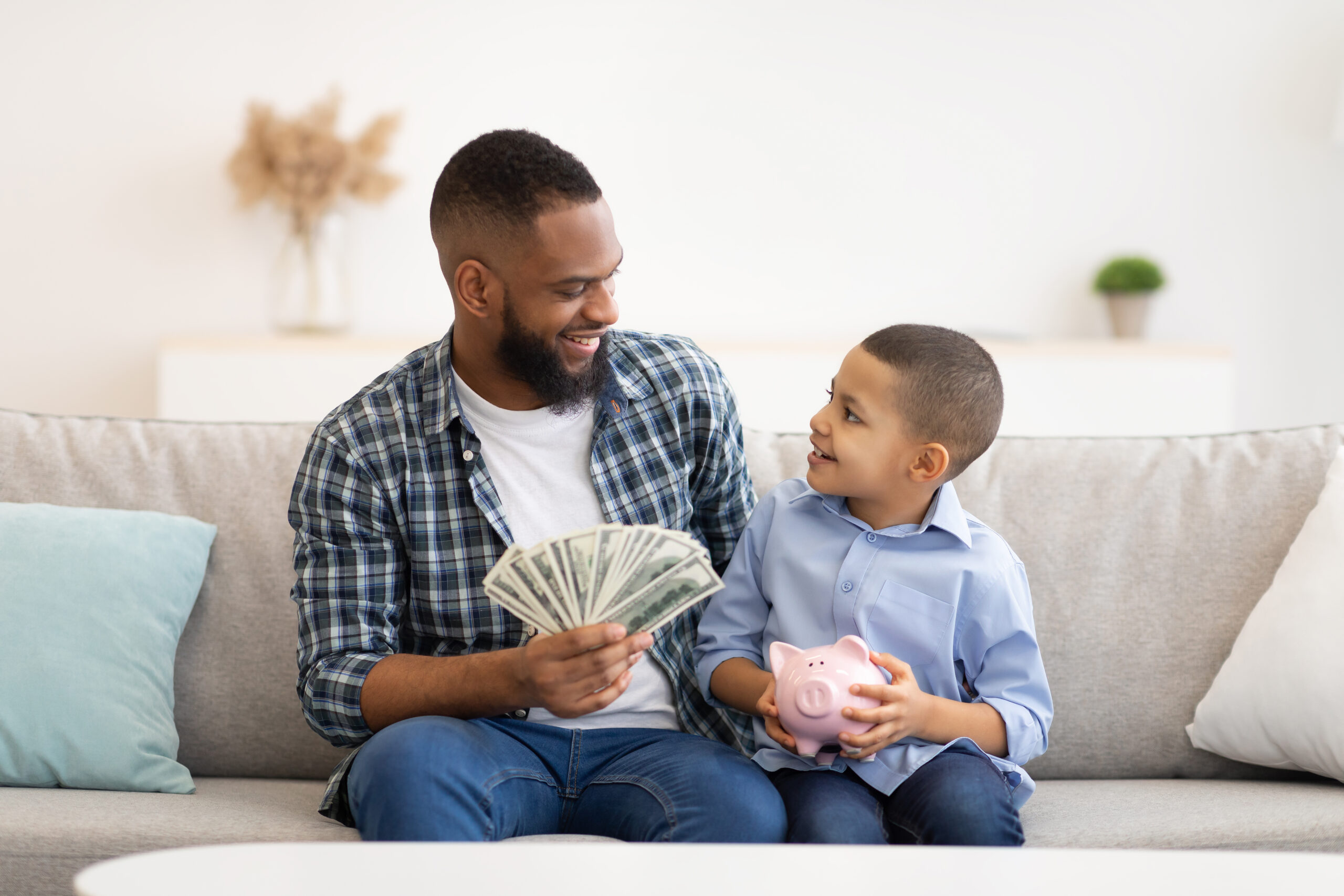 Father and son share a joyful moment discussing money while sitting on a couch, with the son holding a piggy bank.