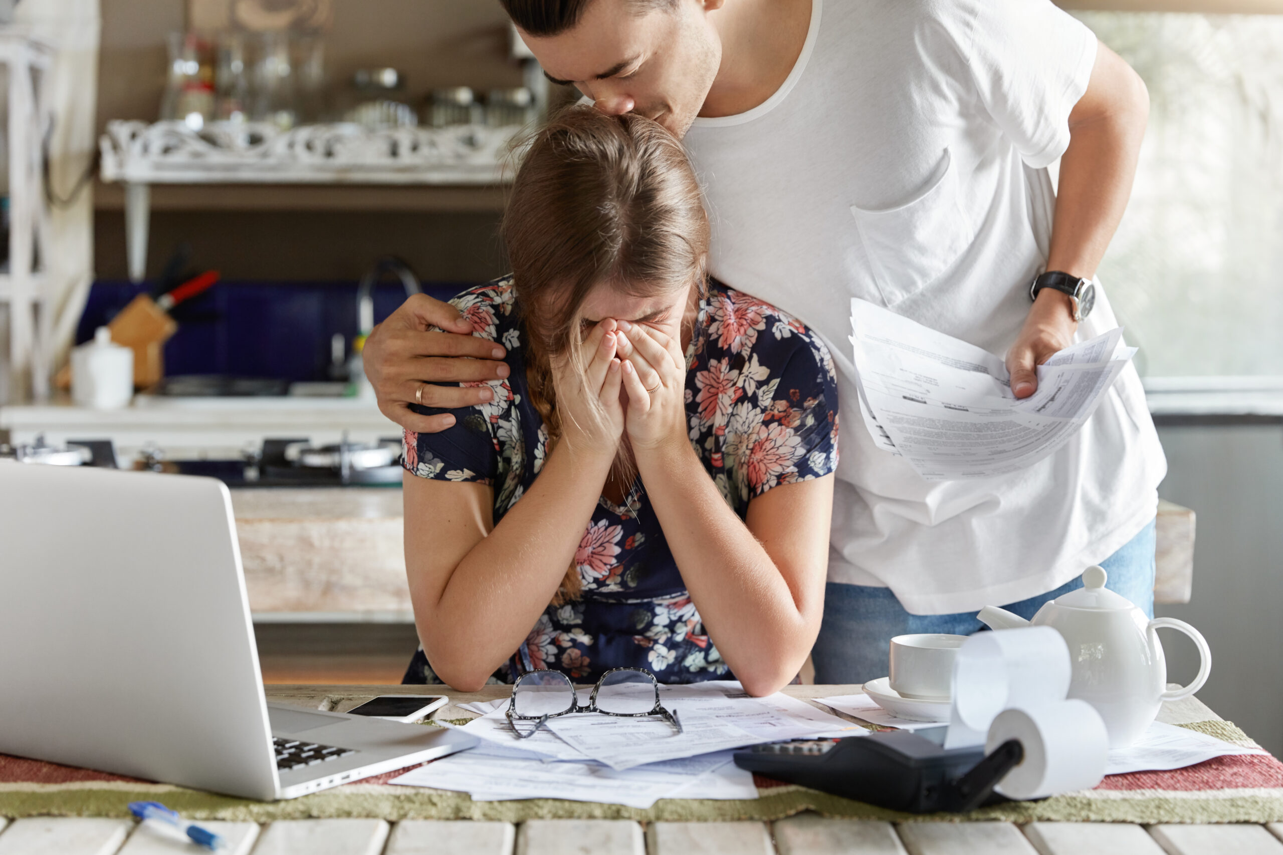 A woman in distress covers her face while a man comforts her, surrounded by paperwork and a laptop on the table.