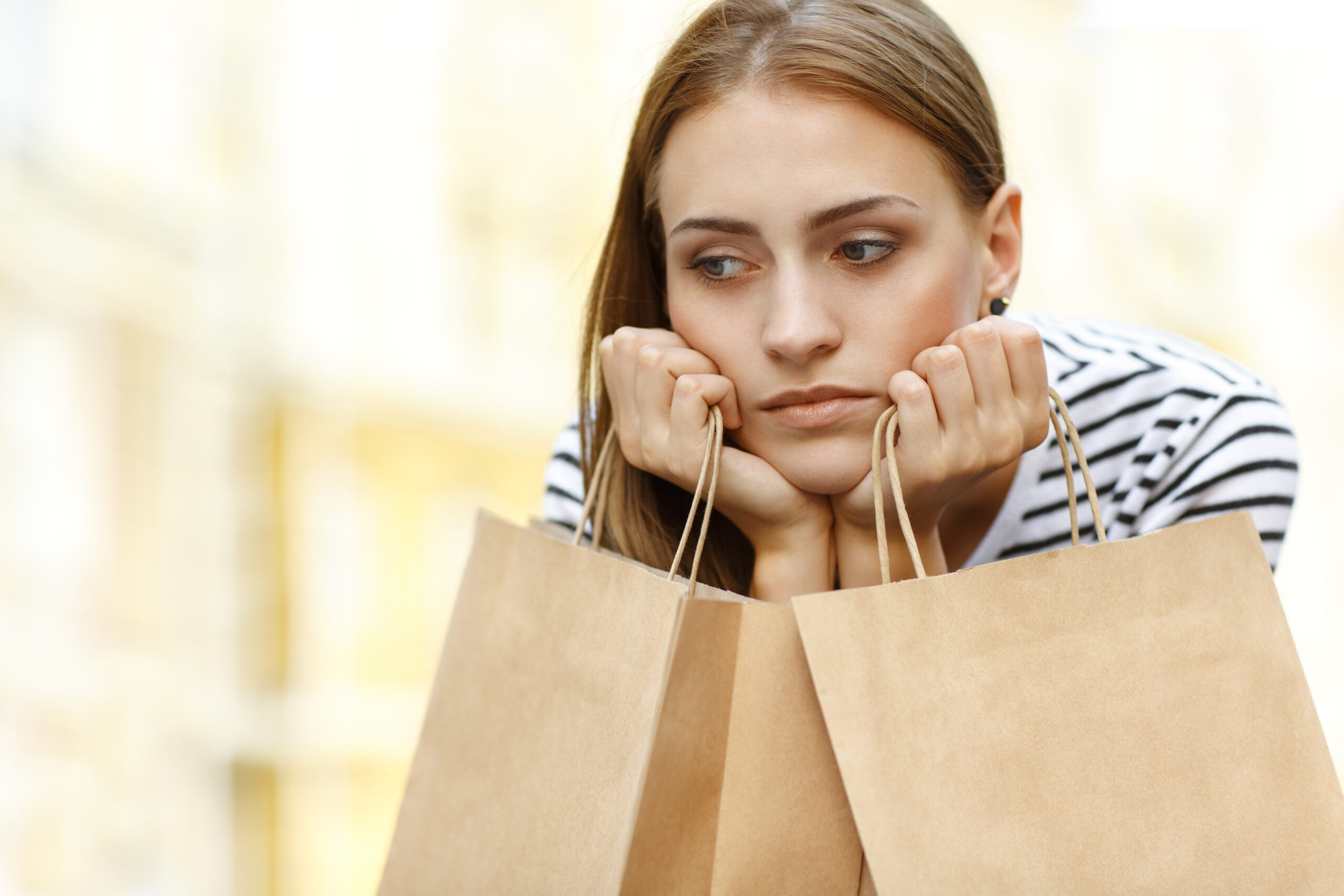 A young woman with a pensive expression rests her chin on her hands while holding shopping bags.