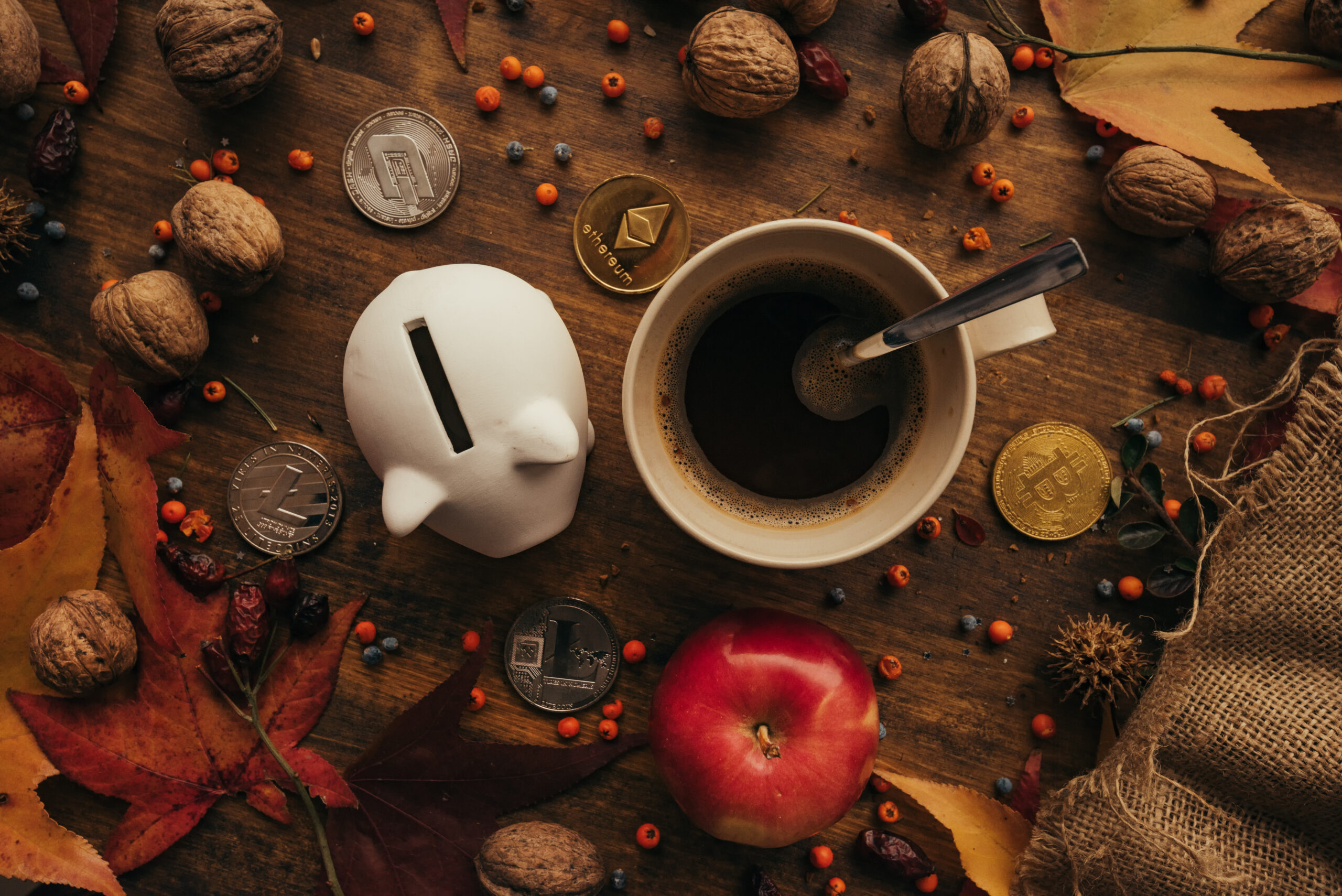 A piggy bank, coffee cup, apple, and autumn leaves surround various coins on a wooden table.