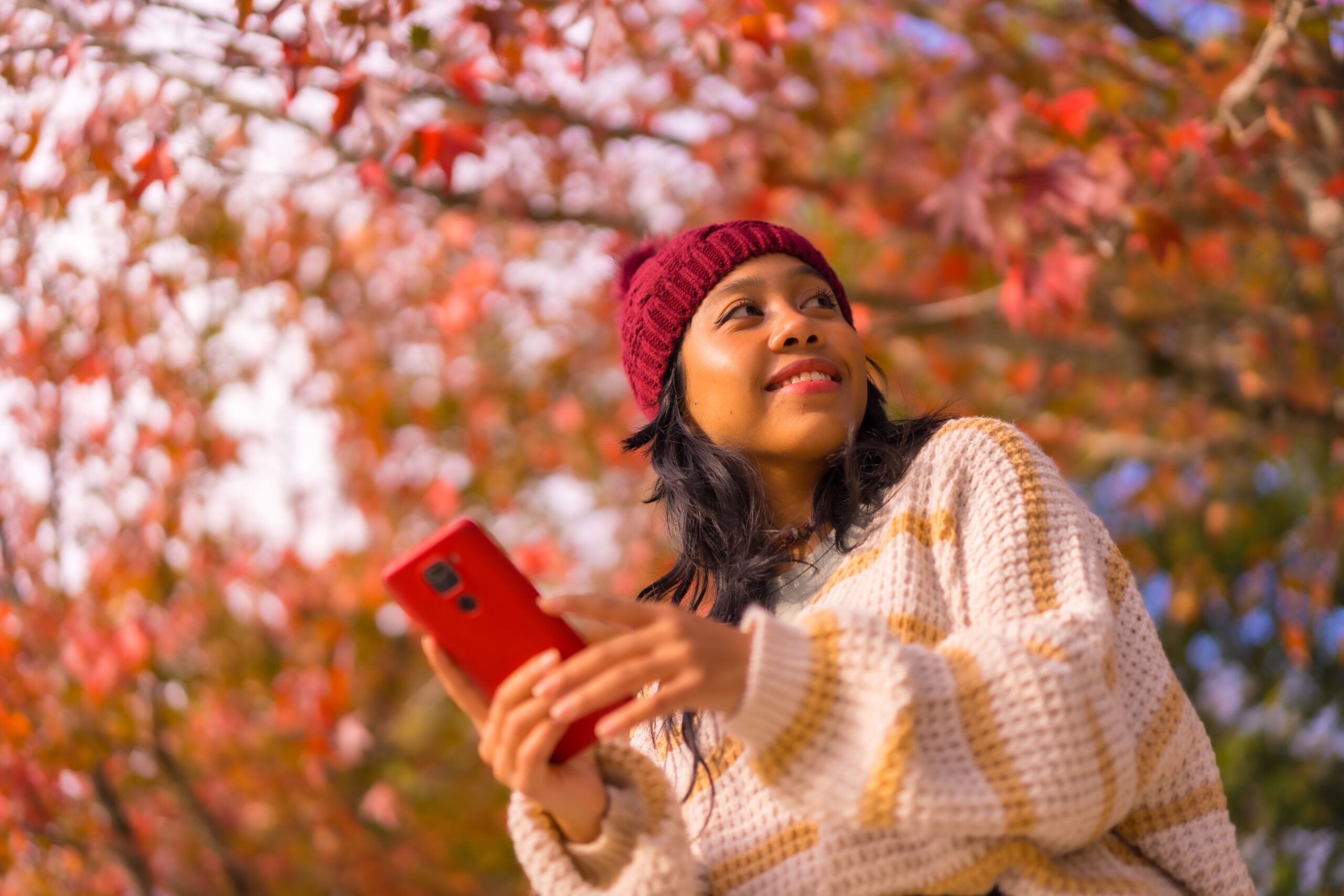 Young woman in a cozy sweater and beanie smiles while holding a red phone amidst vibrant autumn leaves. Fall financial reset