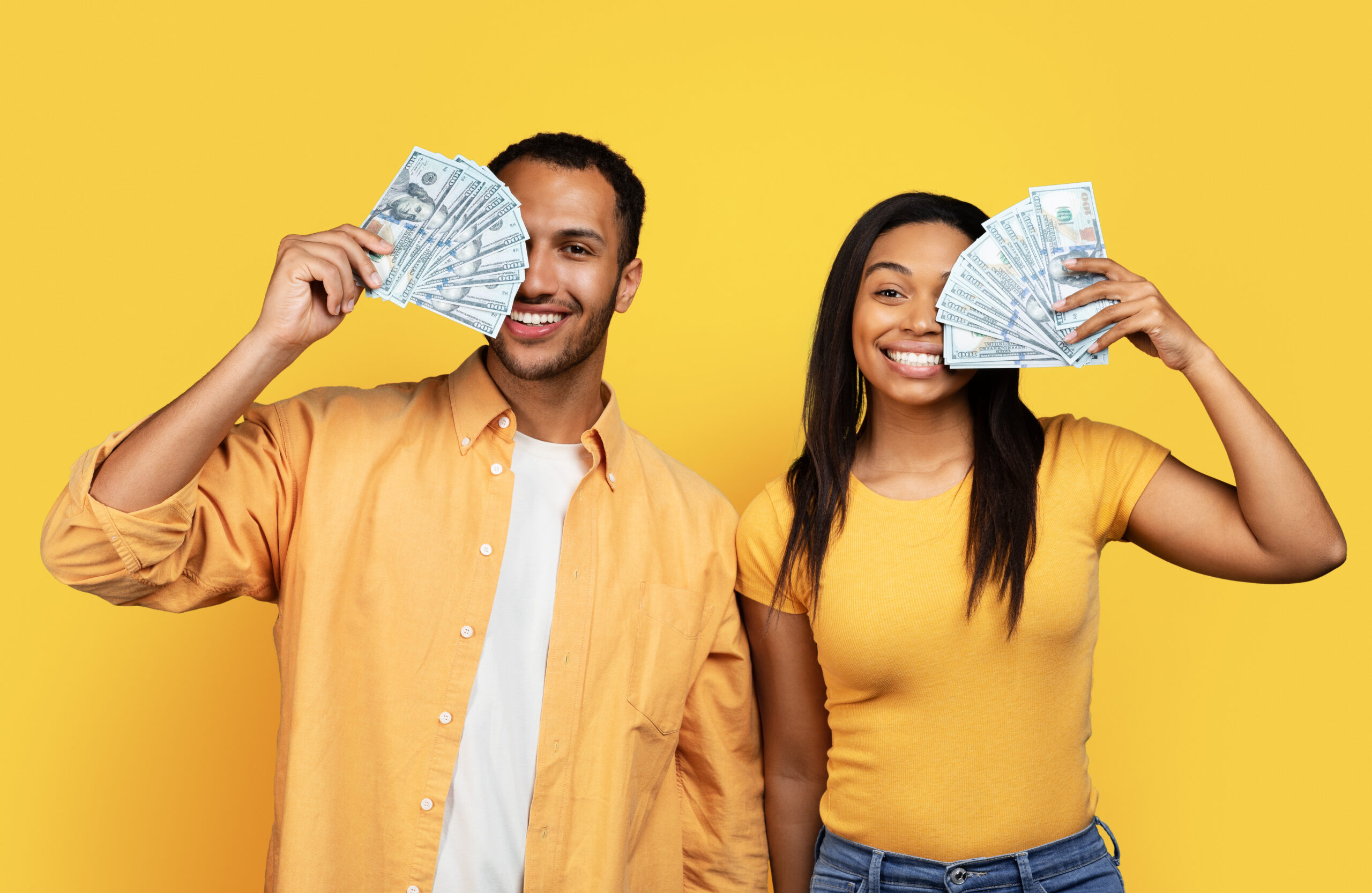 Two smiling individuals hold stacks of cash in front of a bright yellow background.